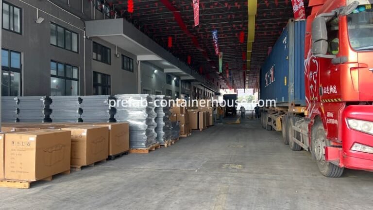 Factory loading zone featuring stacked gray panels and cardboard boxes for portable toilets next to a red delivery truck.
